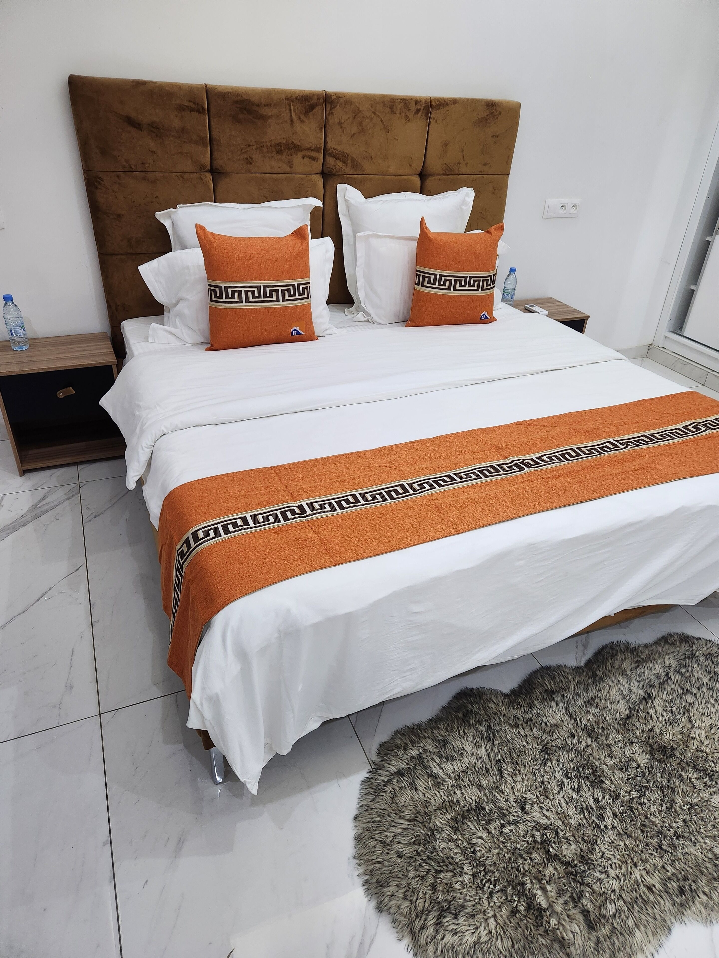 A neatly made bed with white bedding and two orange pillows with a black pattern, a brown upholstered headboard, a small wooden nightstand with a water bottle, and a fluffy rug on the floor.