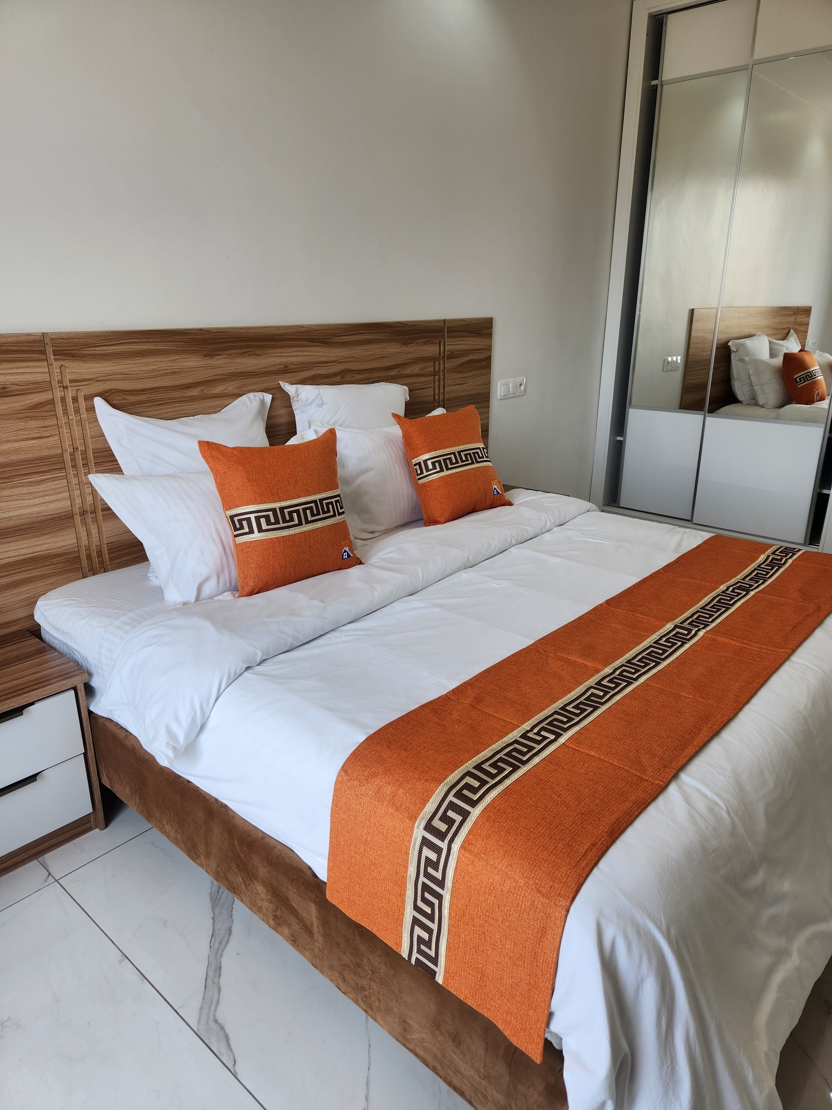 A neatly made bed with white linens and orange pillows, a wooden headboard, and a mirror cabinet to the right.