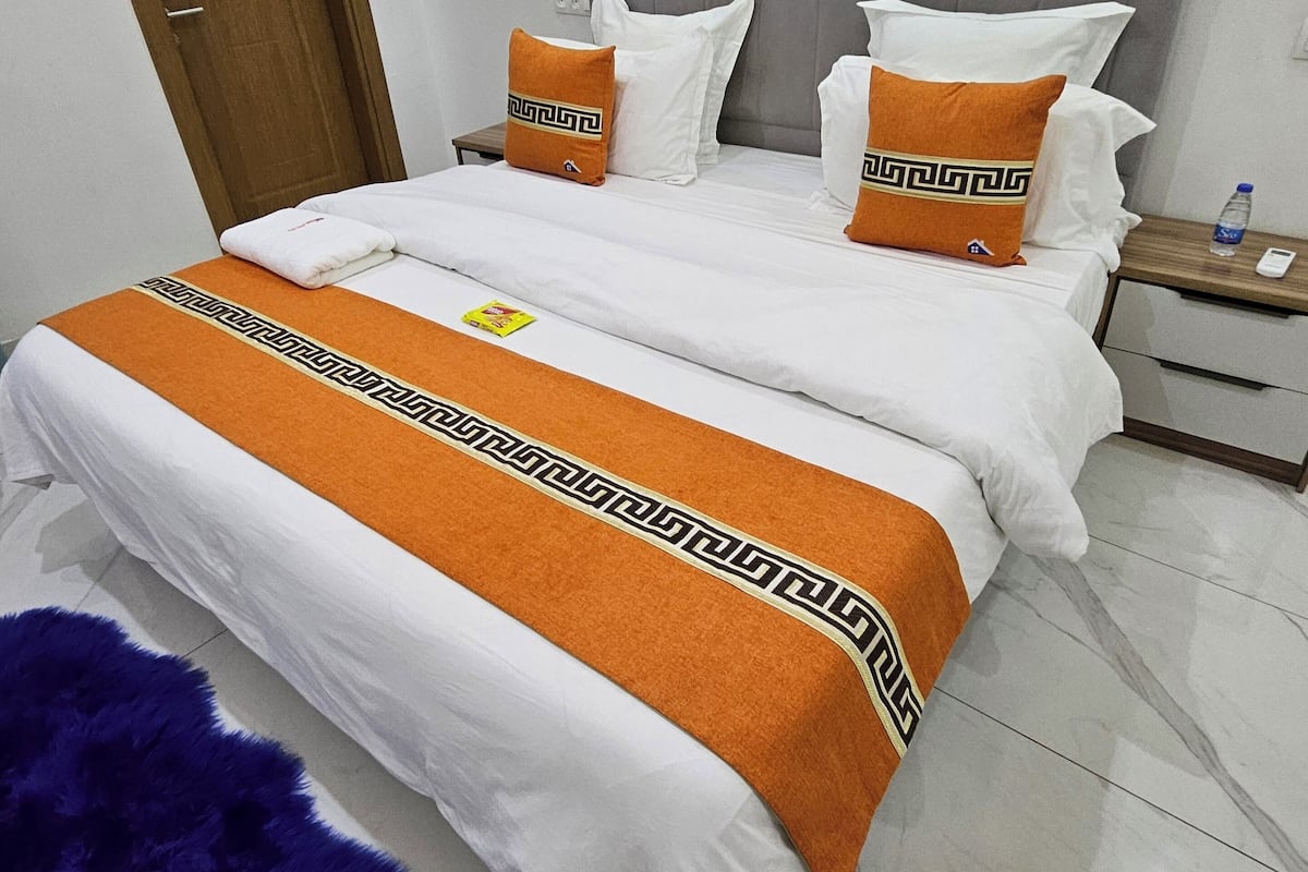 Uma cama de casal com cabeceira cinza, roupa de cama branca e almofadas decorativas laranja.