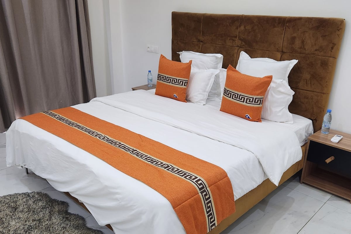 Uma cama bem arrumada com colcha branca e laranja, um tapete fofinho e uma mesinha de cabeceira de madeira com uma garrafa de água e um telefone.