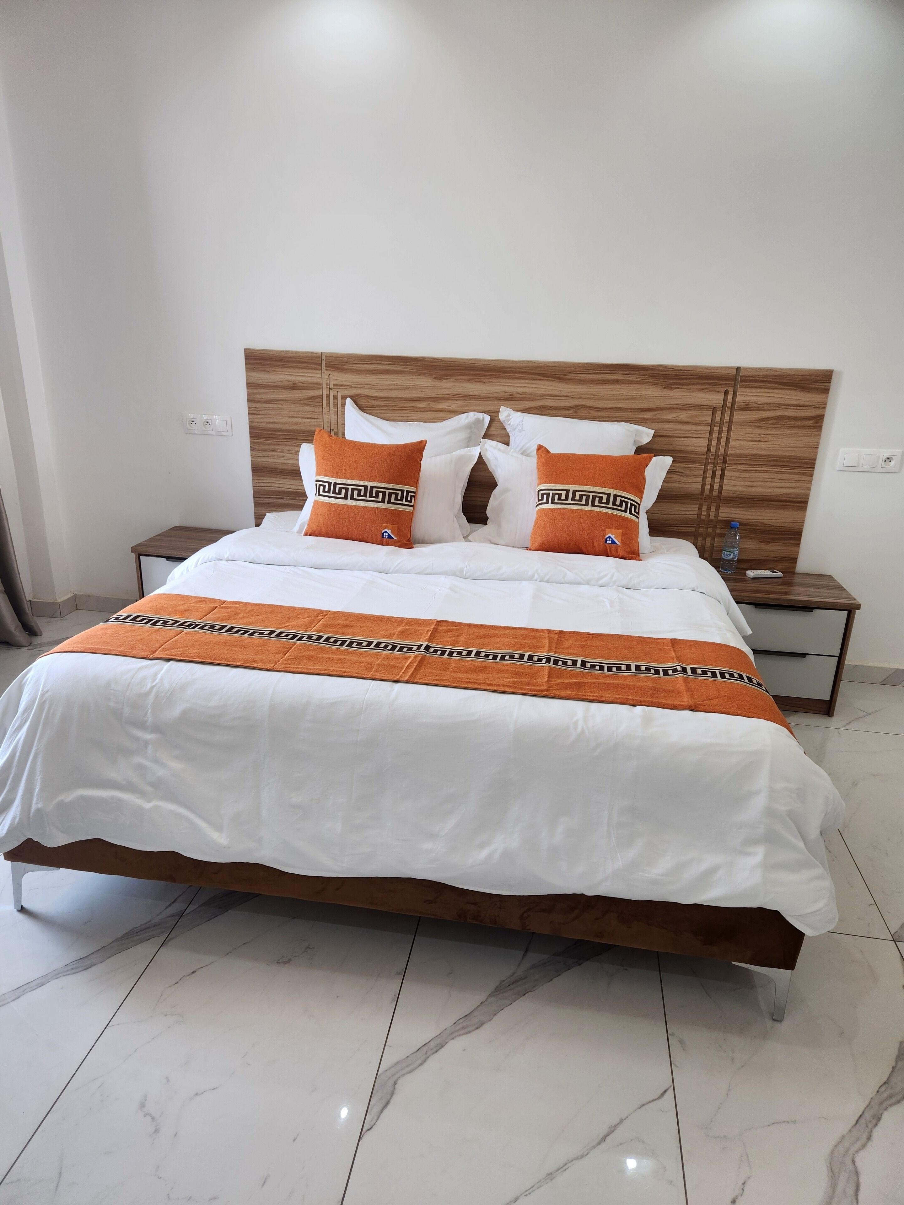 A neatly made bed with white and orange bedding, a wooden headboard, and a marble floor.