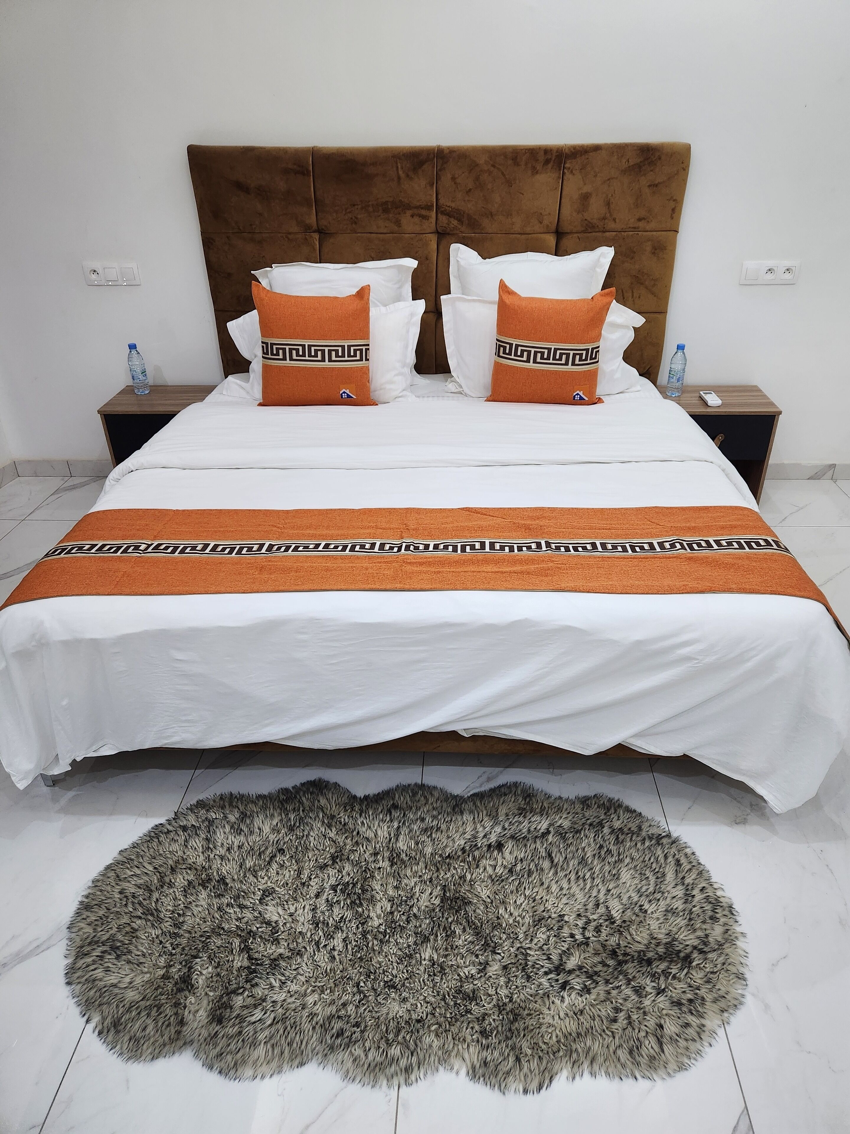 Een net opgemaakt bed met een bruin hoofdeinde, witte en oranje kussens, en een bijpassend oranje plaid. Op de vloer ligt een zacht grijs vloerkleed.