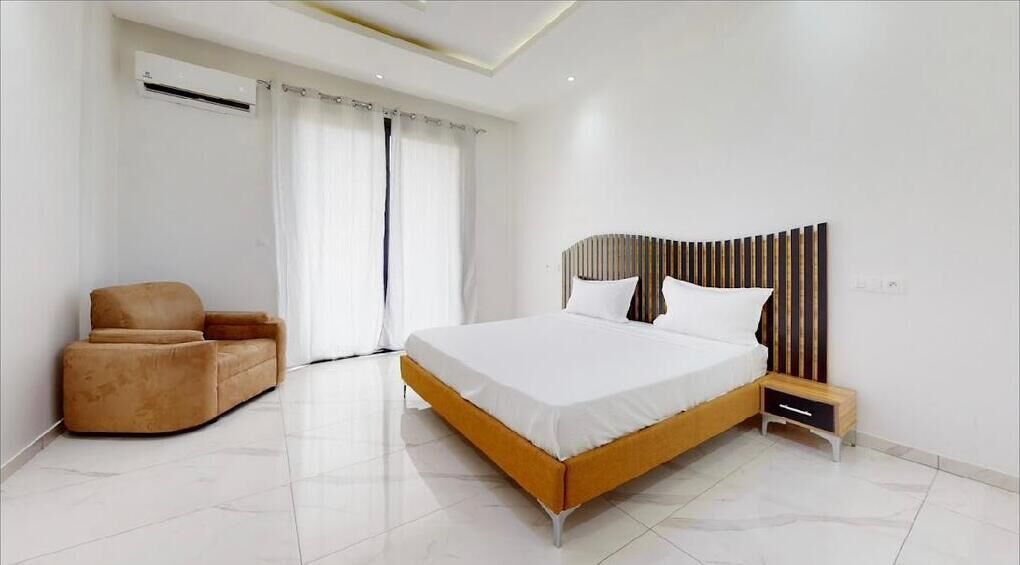 A modern bedroom with a large bed, a small bedside table, a chair, and a window with curtains.