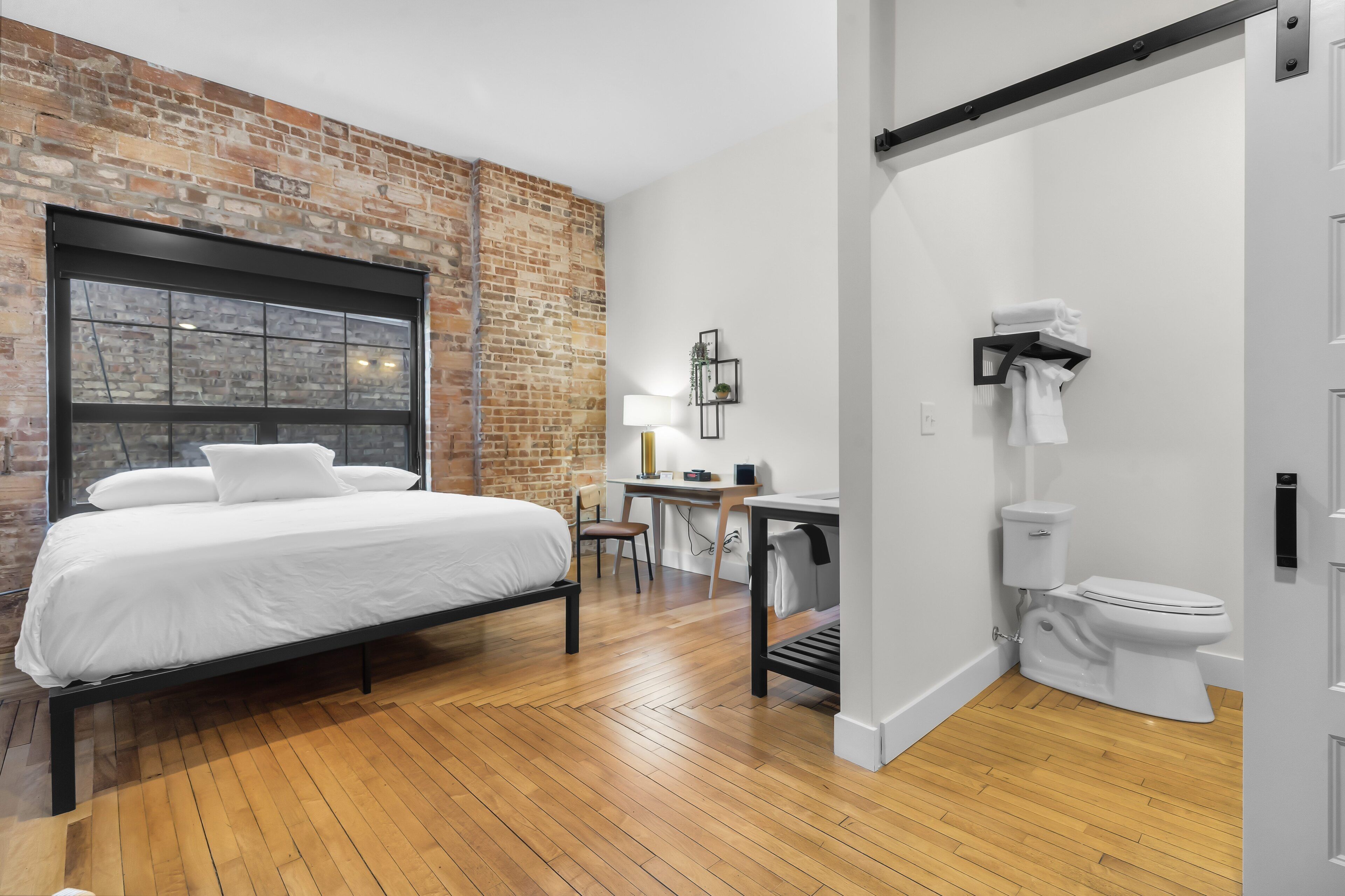 Ein modernes Schlafzimmer mit Backsteinwand, großem Fenster, Bett, Schreibtisch und Toilette.