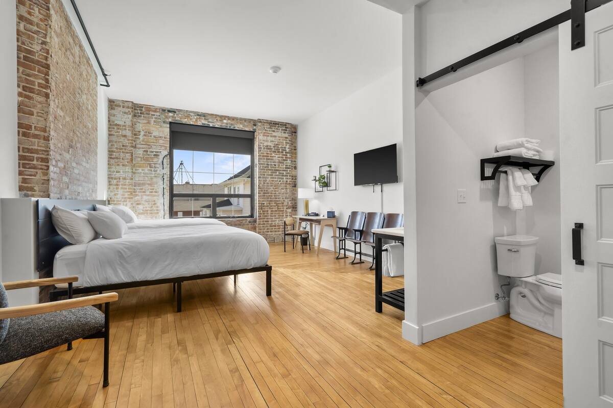 Une chambre moderne avec un lit, un plancher en bois, un mur en briques et un coin bureau avec des chaises.