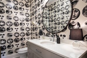 Ein Badezimmer mit einer Wand, die mit Schwarz-Weiß-Porträts versehen ist, einem runden Spiegel, einem weißen Waschbecken und einer Toilette.