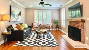 A living room with a fireplace, a ceiling fan, and a large window with blinds. - The Sivley 2BR Classic Home by Velvet Ditch Villas (Oxford)