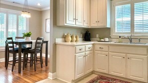 A kitchen with white cabinets, a dining table with chairs, and a window with blinds. - The Sivley 2BR Classic Home by Velvet Ditch Villas (Oxford)