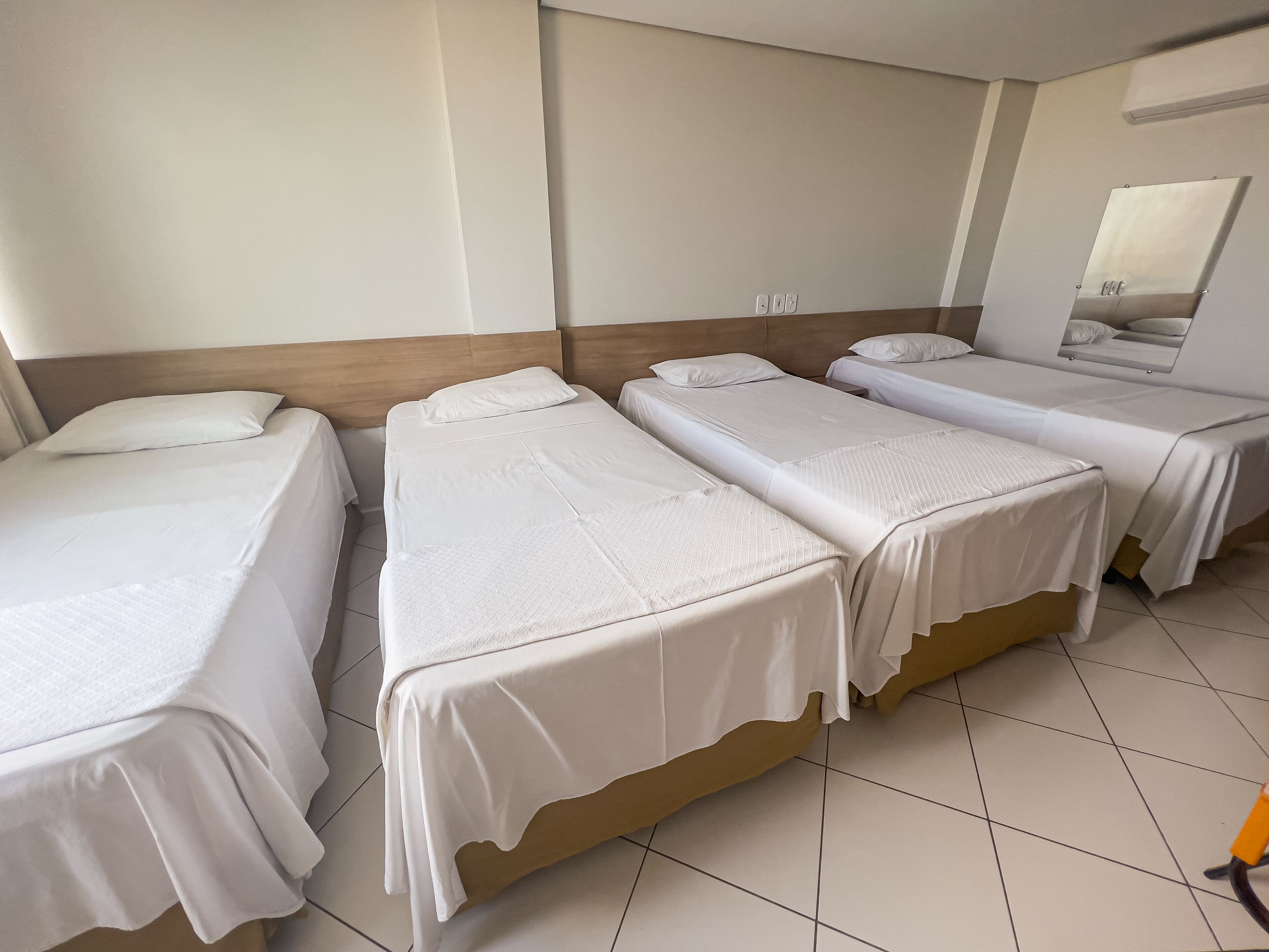 Quarto de hotel com quatro camas de solteiro, cada uma com roupa de cama branca e cabeceira de madeira. O quarto tem piso de azulejos e um espelho grande na parede.