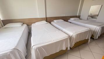 Quarto de hotel com quatro camas de solteiro, cada uma com roupa de cama branca e cabeceira de madeira. O quarto tem piso de azulejos e um espelho grande na parede.