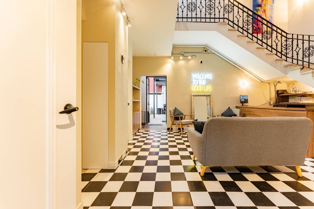 Un vestíbulo de hotel con piso de cuadros, una escalera con barandilla de hierro forjado y un letrero de neón que dice "WELCOME TO THE GOOD LIFE".
