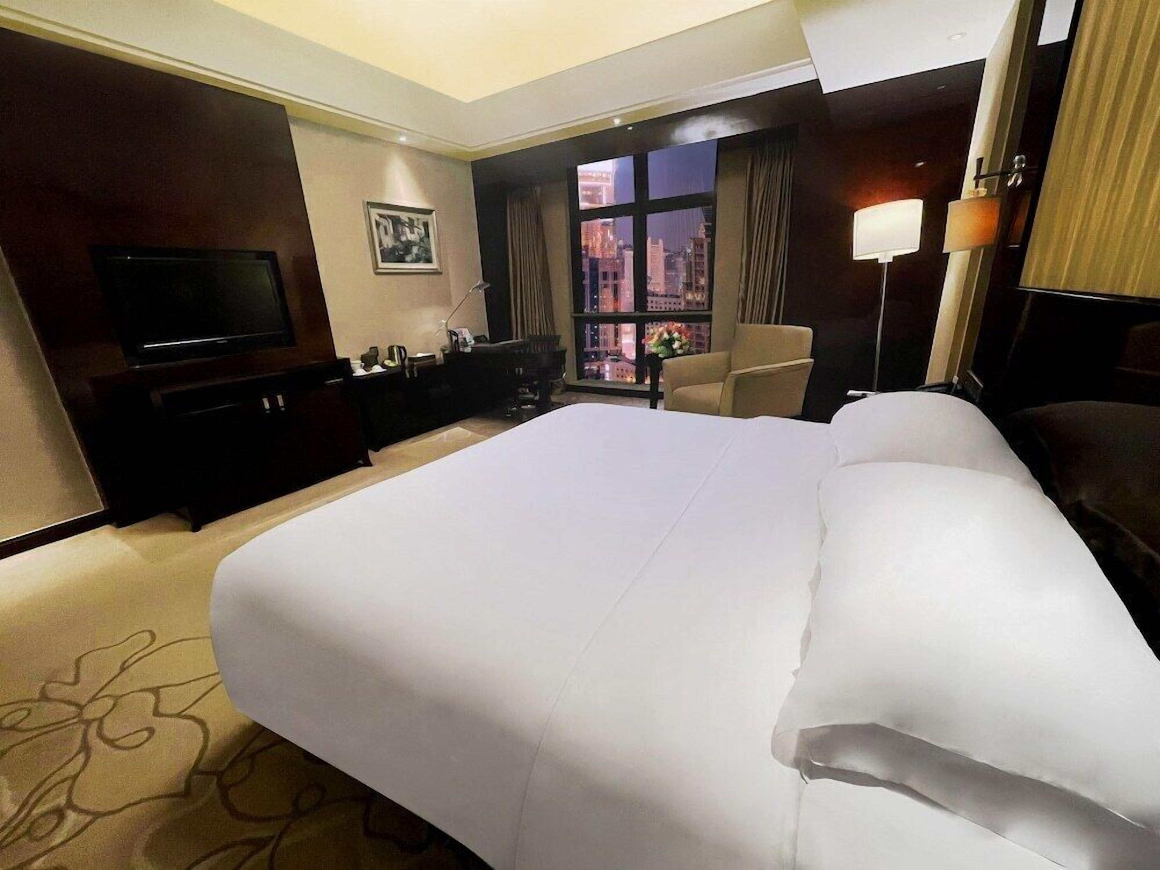 A hotel room with a large bed, a television, a desk, and a window with a city view.