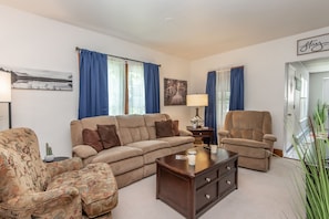 A living room with a beige sofa, patterned armchair, and a brown recliner. There is a wooden coffee table, a side table with a lamp, and a painting on the wall.