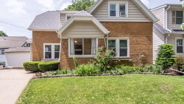 A two-story house with a brick facade and white trim, a front porch, and a well-maintained lawn. - 4BD House, Your Central Suburban Oasis, City Lights, Quiet Nights (Wauwatosa)