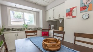 A dining area with a wooden table, chairs, a blue placemat, a wooden bowl, a window with blinds, a kitchen area with cabinets, a coffee maker, and wall art. - 4BD House, Your Central Suburban Oasis, City Lights, Quiet Nights (Wauwatosa)