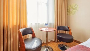 Une chambre d’hôtel avec deux fauteuils en cuir, une petite table ronde avec une bouteille d’eau et un lit recouvert d’une couverture rouge.
