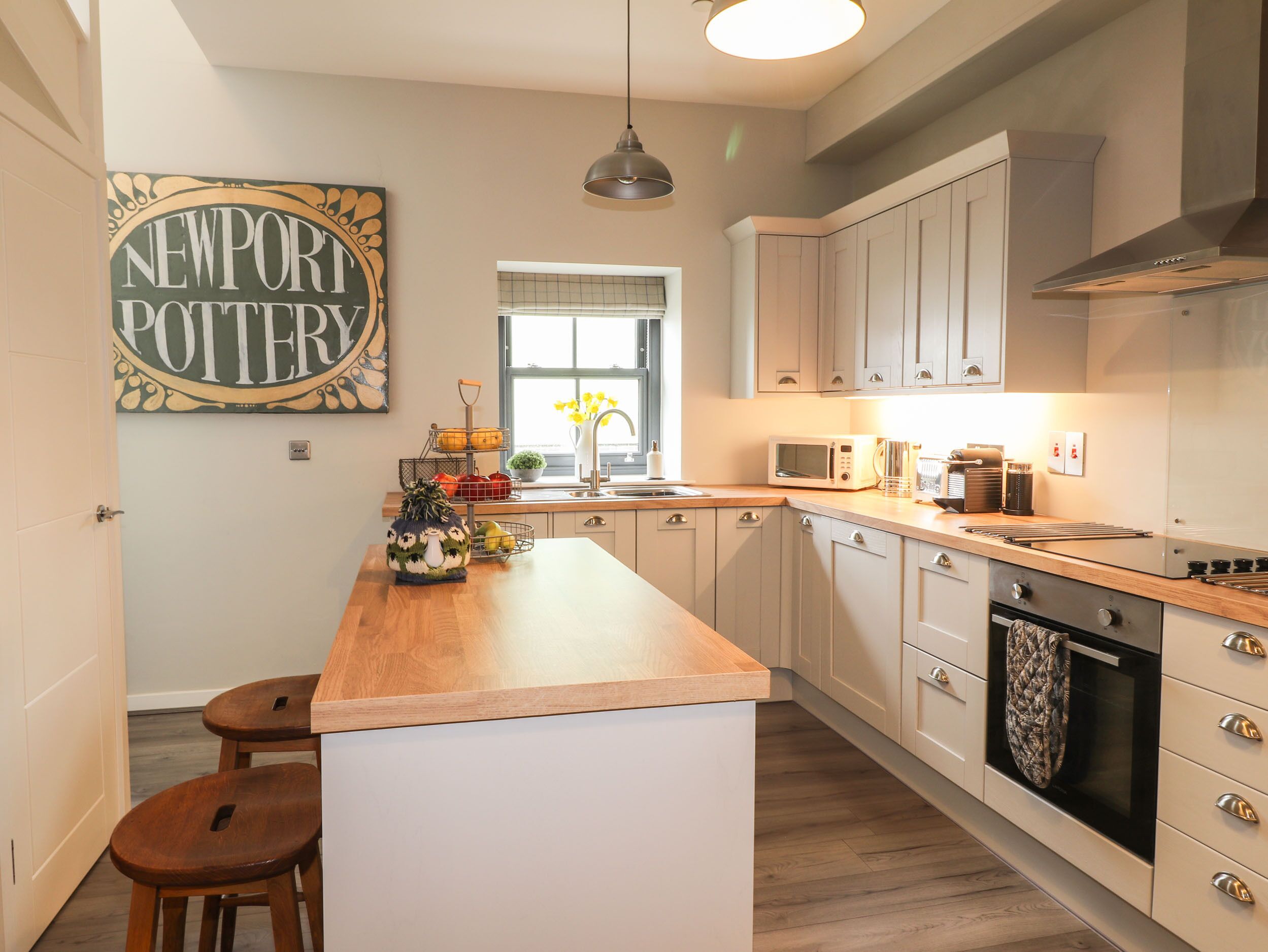 A modern kitchen with a central island, wooden stools, a hanging light fixture, and a 'Newport Pottery' sign on the wall.