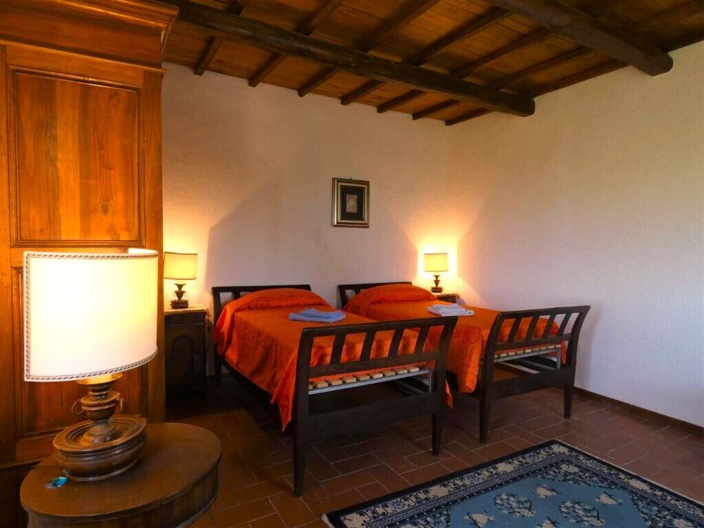 A room with two wooden beds, each with an orange bedspread, a round table with a lamp, and a blue patterned rug on the floor.