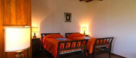 A room with two wooden beds, each with an orange bedspread, a round table with a lamp, and a blue patterned rug on the floor.