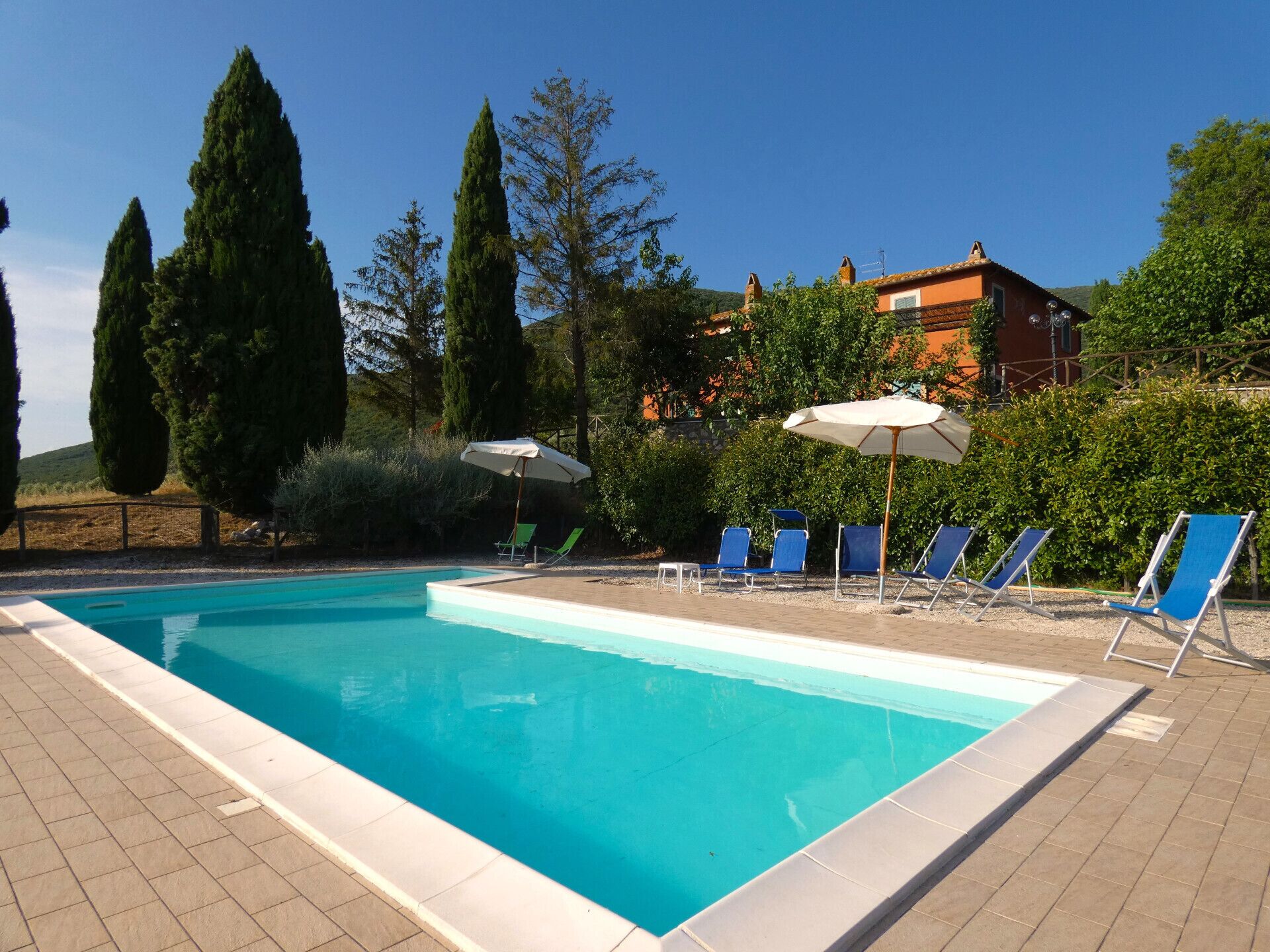 Ein rechteckiger Swimmingpool mit klarem, blauem Wasser, umgeben von einer gefliesten Liegefläche. Mehrere Liegestühle mit blauen Kissen und weißen Sonnenschirmen sind in Reihen angeordnet.