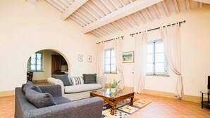 A living room with a sofa, armchairs, a coffee table, and a rug. There are two windows with curtains, a door, and a kitchen visible in the background.