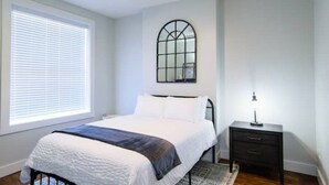 A bedroom with a bed, a nightstand, a window with blinds, a ceiling fan, and a wall-mounted mirror.