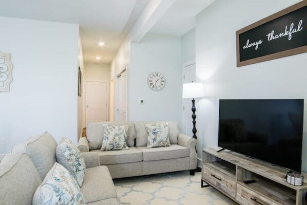 Ein Wohnzimmer mit einem beigefarbenen Sofa, einem Flachbildfernseher auf einem Holzbodenständer, einer Wanduhr und einem Schild mit der Aufschrift 'always thankful'.