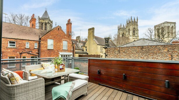 Una terraza en la azotea con mobiliario de mimbre, una mesa con un florero y vistas a edificios históricos al fondo.