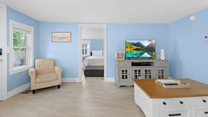 Una habitación con pared azul claro, un sillón color beige, una mesa de centro de madera, un televisor y una cama al fondo.