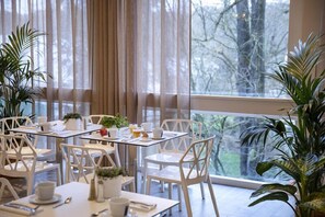 A dining area with white chairs and tables, large windows with curtains, and potted plants. - Hotel du Port (Nogent-sur-Marne)