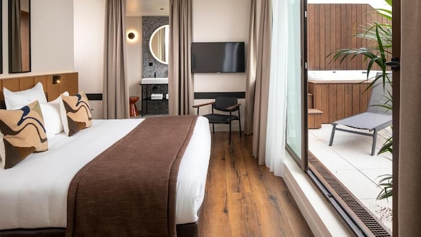 A hotel room with a large bed, a TV, a small desk, and a view of a wooden deck and outdoor area. - Hotel du Port (Nogent-sur-Marne)