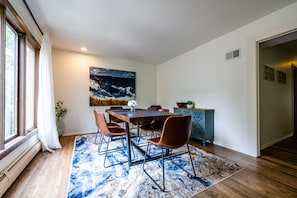 A dining area with a wooden table, four chairs, a blue and white patterned rug, a painting on the wall, and a cabinet with decorative items. - Spacious 4BR Retreat • Private Beach • Heated Outdoor Pool is OPEN • Golf Cart (Coloma)