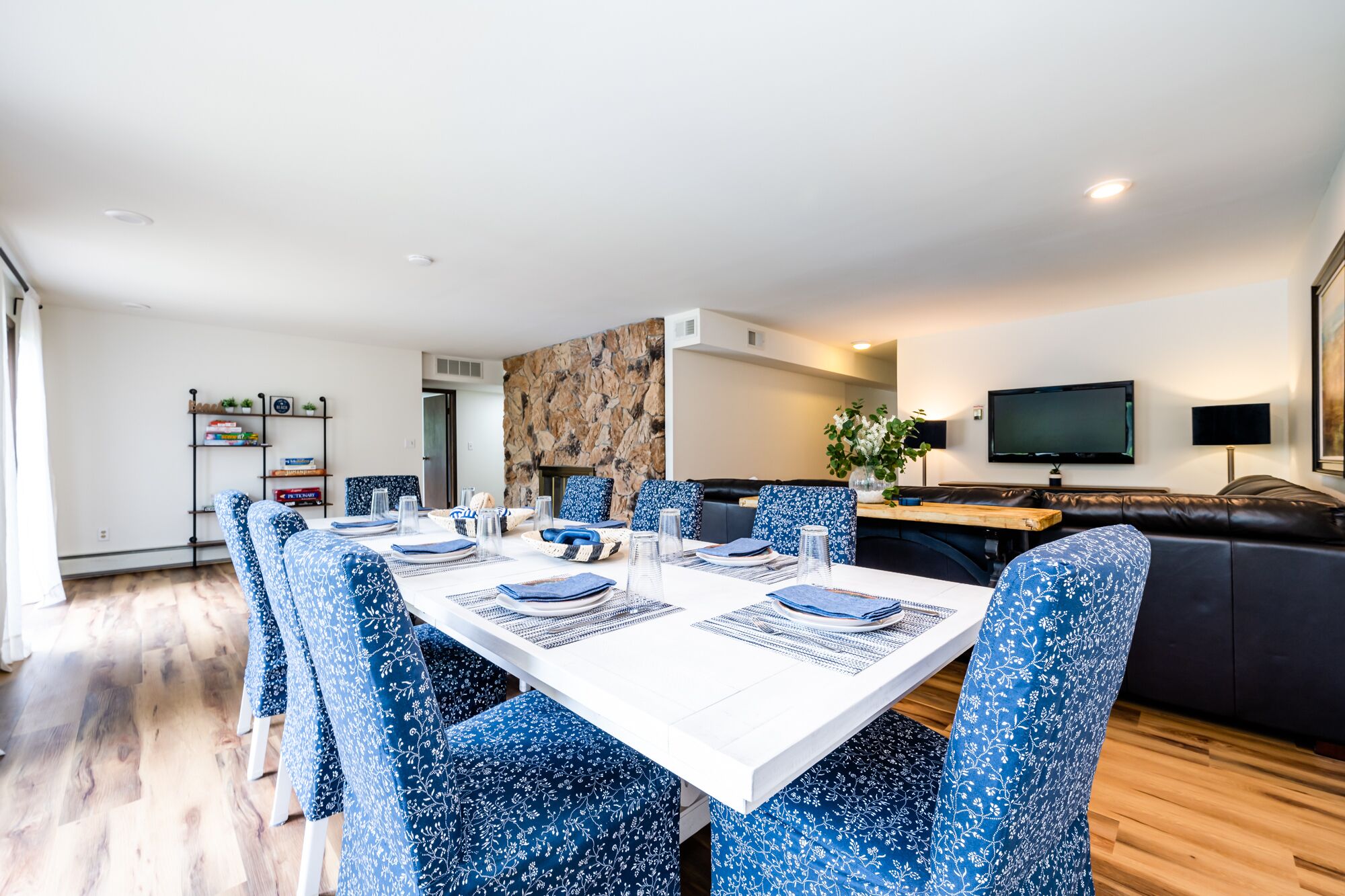 Un coin repas avec une table blanche dressée pour quatre personnes, des chaises à motifs bleus, un mur décoratif en pierre, un canapé en cuir noir et un téléviseur à écran plat fixé au mur.
