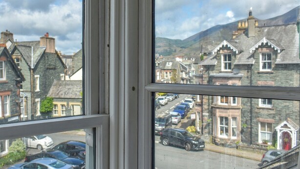 A view through a window of a street with parked cars, stone buildings, and a mountain in the background. - Enjoy a family holiday right here in the heart of the Lake District. (Keswick)