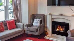 A living room with a grey armchair, a red and grey sofa, a fireplace, and a television mounted above it. - Enjoy a family holiday right here in the heart of the Lake District. (Keswick)