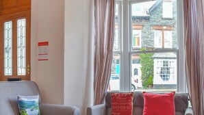 A cozy living room with a grey sofa set, a red pillow with a home-themed message, and a window with curtains. - Enjoy a family holiday right here in the heart of the Lake District. (Keswick)