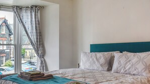 A neatly made bed with a teal headboard, a window with a view of parked cars and buildings, and a curtain partially drawn. - Enjoy a family holiday right here in the heart of the Lake District. (Keswick)