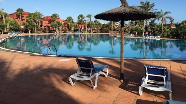 A swimming pool with lounge chairs and umbrellas, surrounded by palm trees and buildings. - Holiday flat in a beautiful complex with large swimming pool and private terrace (Corralejo)