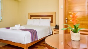 A hotel room with a wooden bed, white bedding, a bedside table with a phone, and a small potted plant.