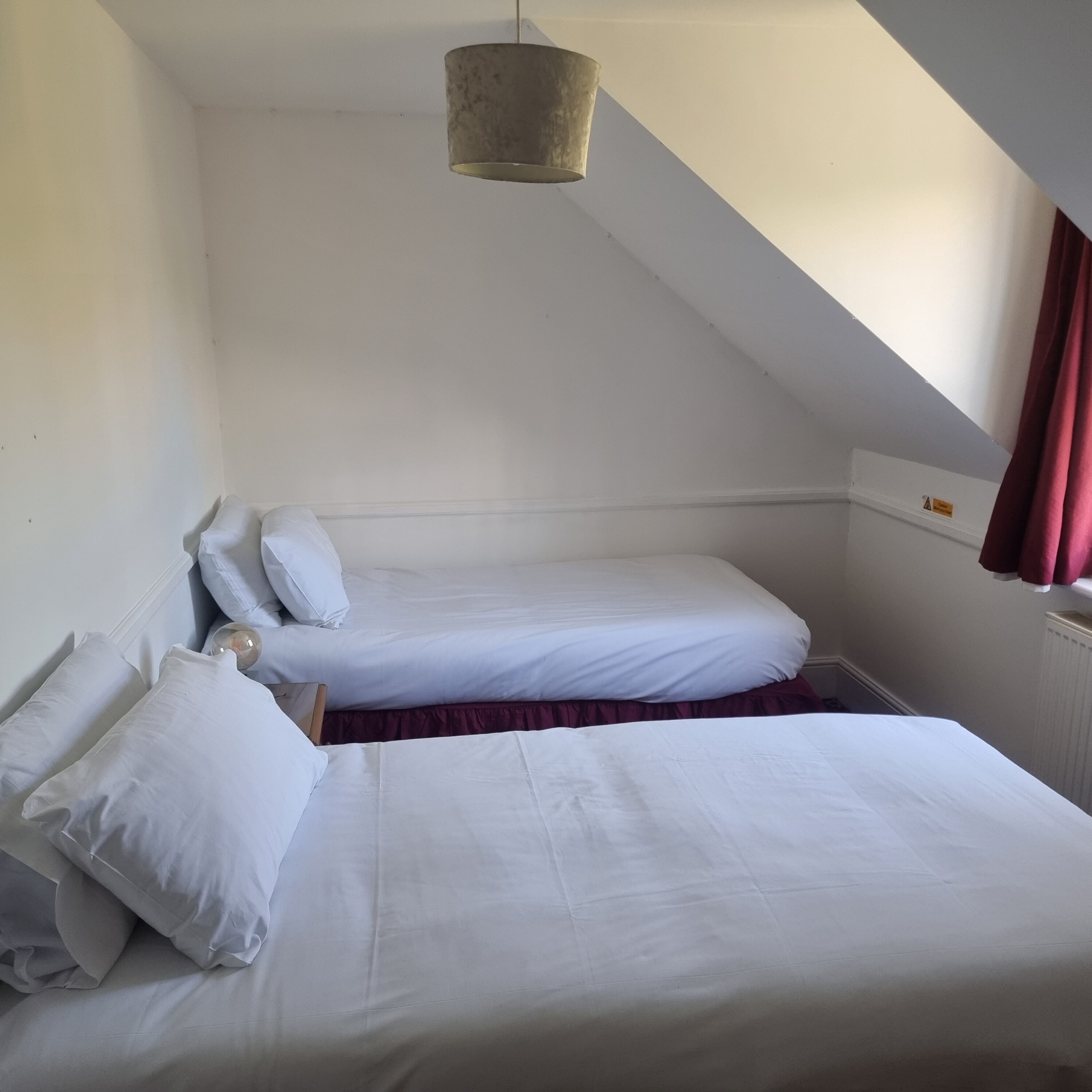 A small, single-room hotel bedroom with two beds, a hanging light fixture, and a window with curtains.