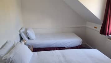 A small, single-room hotel bedroom with two beds, a hanging light fixture, and a window with curtains.