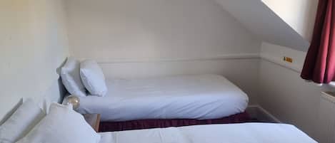 A small, single-room hotel bedroom with two beds, a hanging light fixture, and a window with curtains.