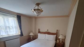Un dormitorio con cama, ventana con cortinas, radiador y mesita de noche con lámpara.