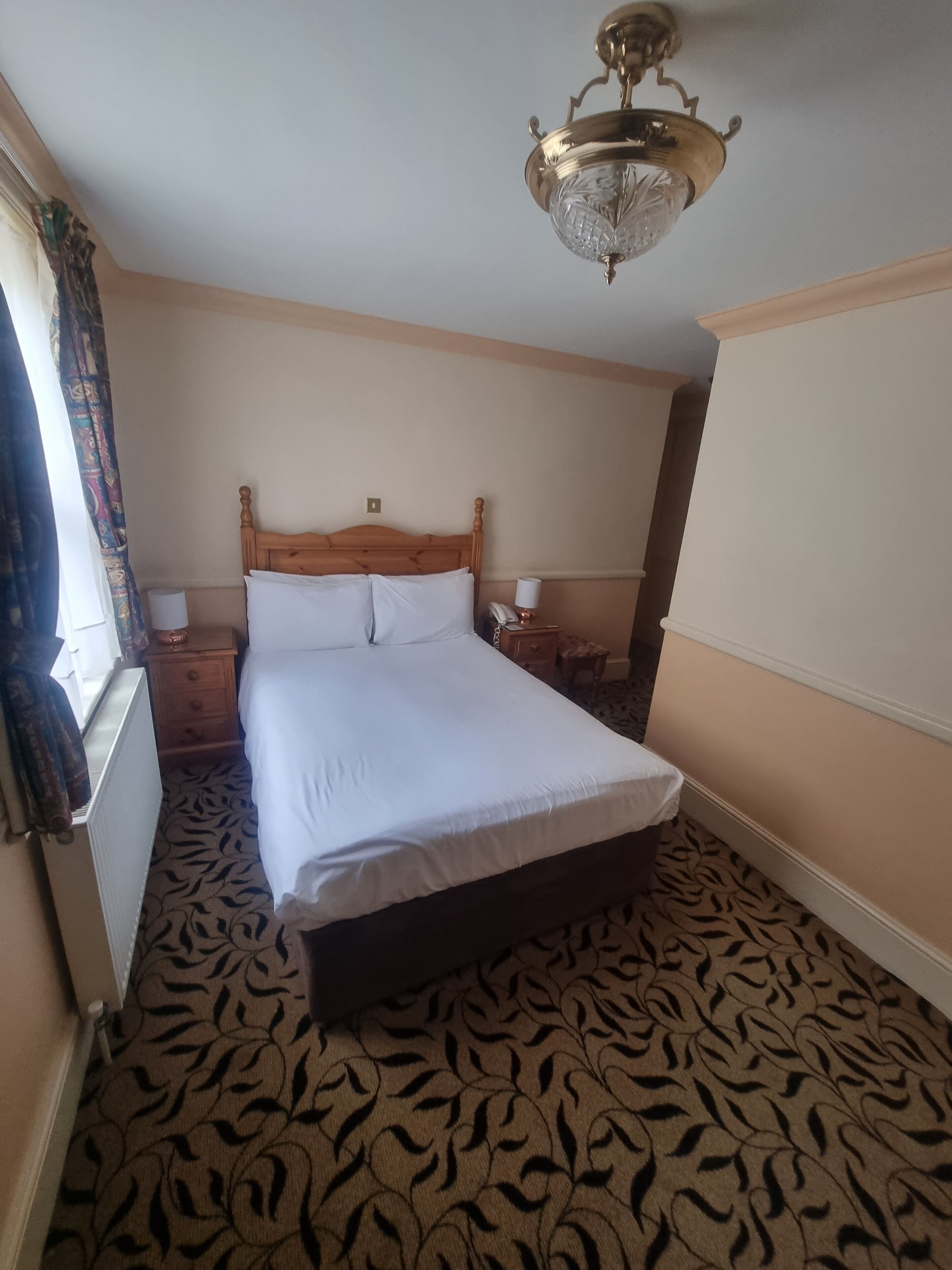 A hotel room with a wooden bed, a patterned carpet, a chandelier, and a window with curtains.