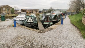 A canal-side boat moored at a dock with a gravel path leading to a grassy area. - The Bike Shed (High Peak)