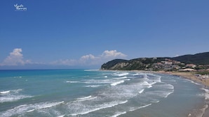 Une plage côtière aux eaux bleues et limpides, bordée d’un rivage sablonneux et d’une colline parsemée de bâtiments.