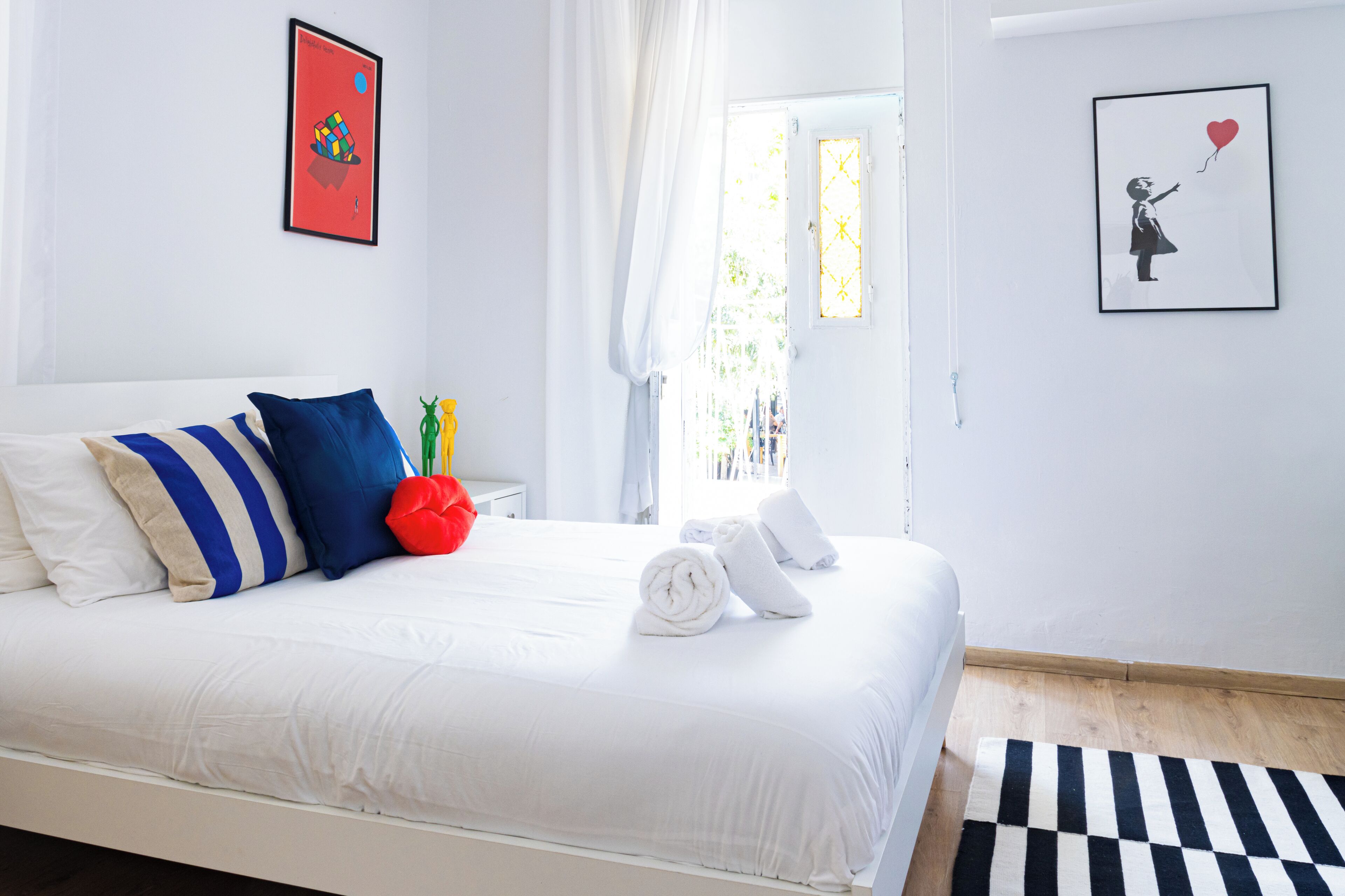 A neatly made bed with white bedding, blue and beige pillows, and a red decorative pillow. There are two framed artworks on the walls, a window with curtains, and a black and white striped rug on the floor.