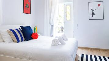 A neatly made bed with white bedding, blue and beige pillows, and a red decorative pillow. There are two framed artworks on the walls, a window with curtains, and a black and white striped rug on the floor.