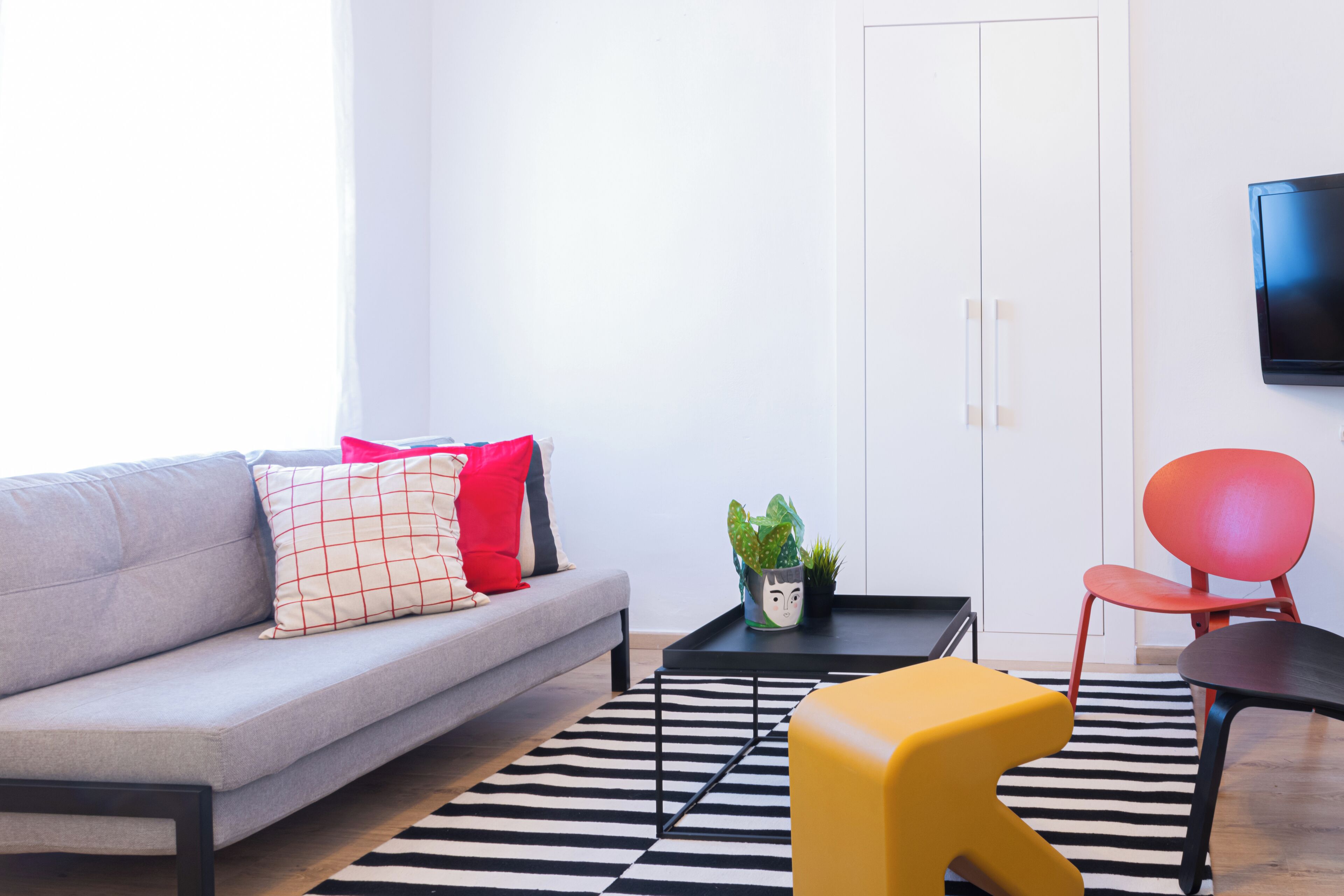 A modern living room with a grey sofa, a black coffee table, a yellow ottoman, a red chair, and a black and white striped rug.