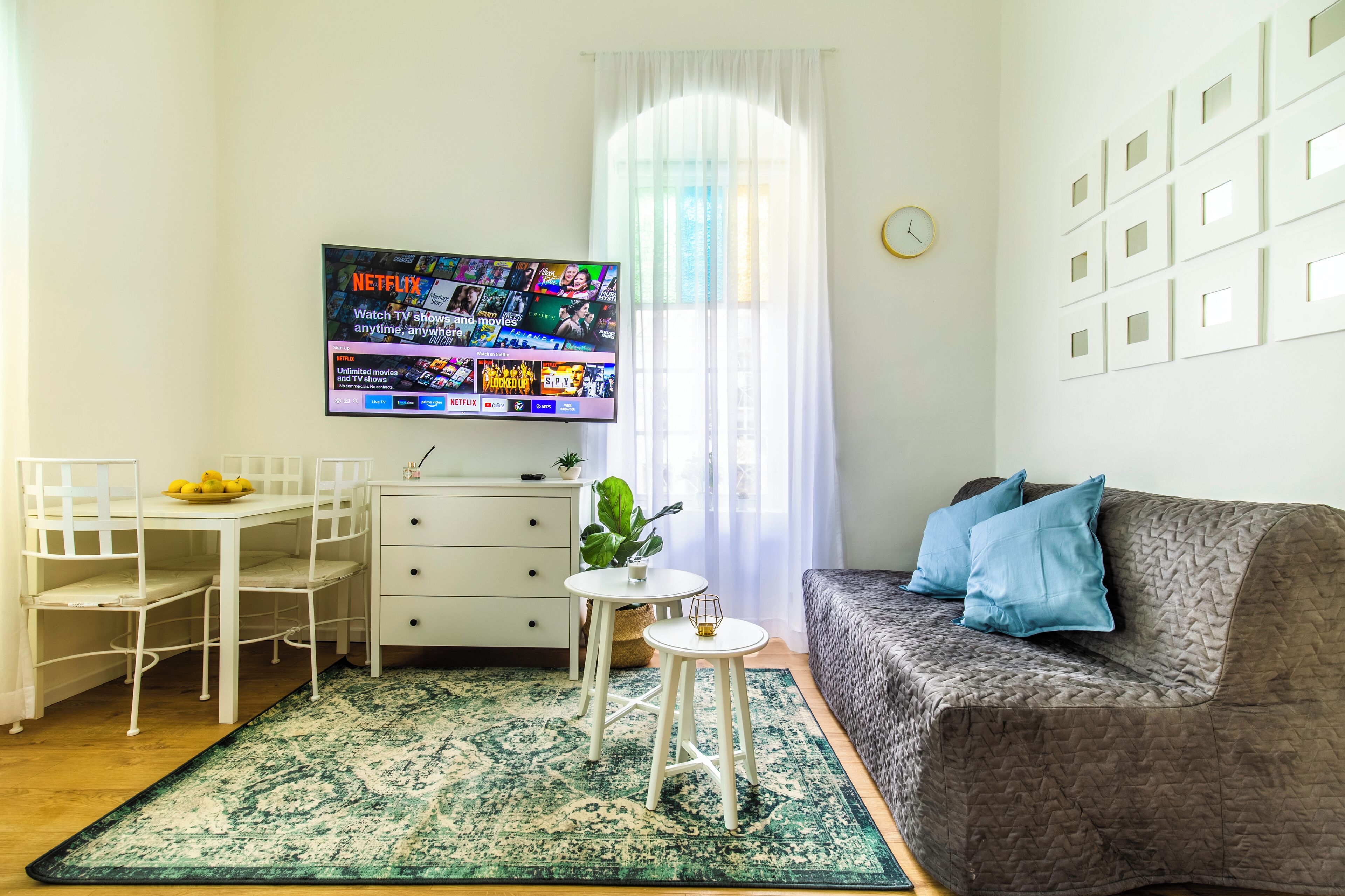 Uma sala de estar com televisão de tela plana exibindo conteúdo da Netflix, uma mesa de jantar branca com cadeiras, uma pequena mesa redonda com uma planta, um sofá cinza com almofadas azuis e um tapete estampado.